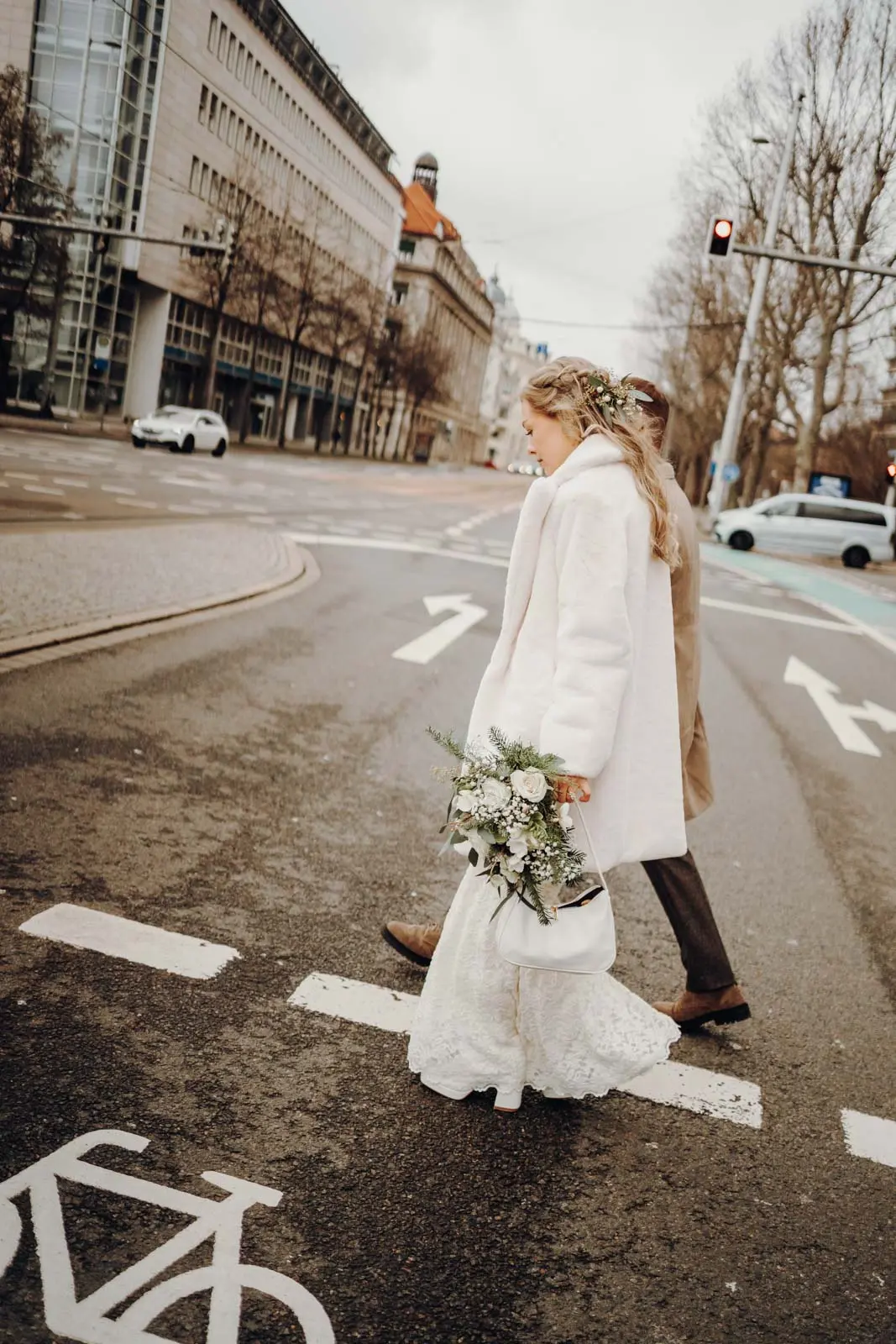 Braut ueberquert Strasse in Leipzig mit Winterstrauss Braut im Spitzenkleid und weissem Mantel ueberquert einen Zebrastreifen in der Leipziger Innenstadt, Winterstrauss in der Hand.