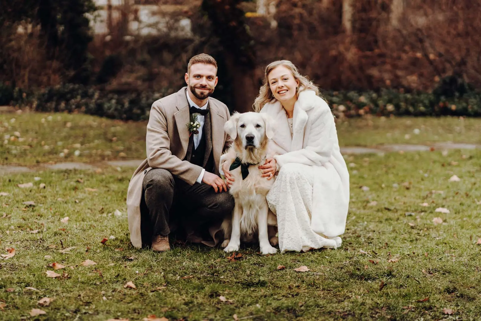 Brautpaar mit Golden Retriever nach der Trauung Braut und Braeutigam kauern gemeinsam mit ihrem weissen Golden Retriever auf dem Rasen im Hochzeitsoutfit.