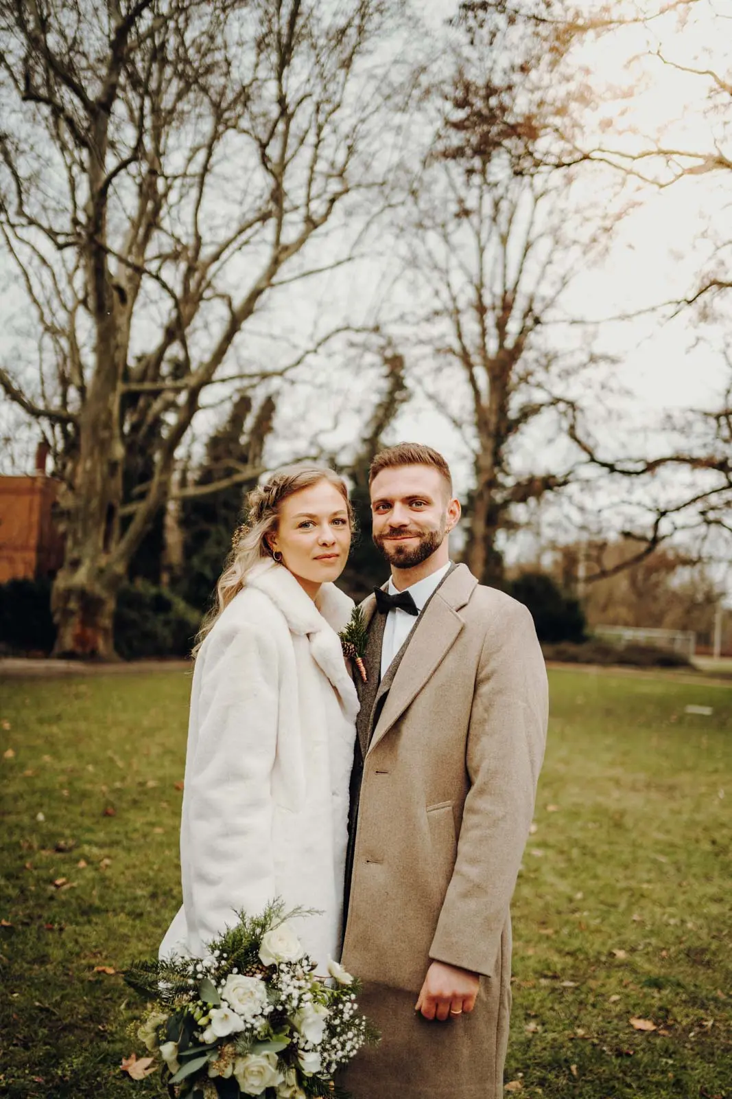 Brautpaarportrait im Winterpark – Frontale Aufnahme Brautpaar steht Seite an Seite im winterlichen Park und blickt in die Kamera, Braut haelt einen Winterstrauss. Hochzeit Ratsplenarsaal Leipzig