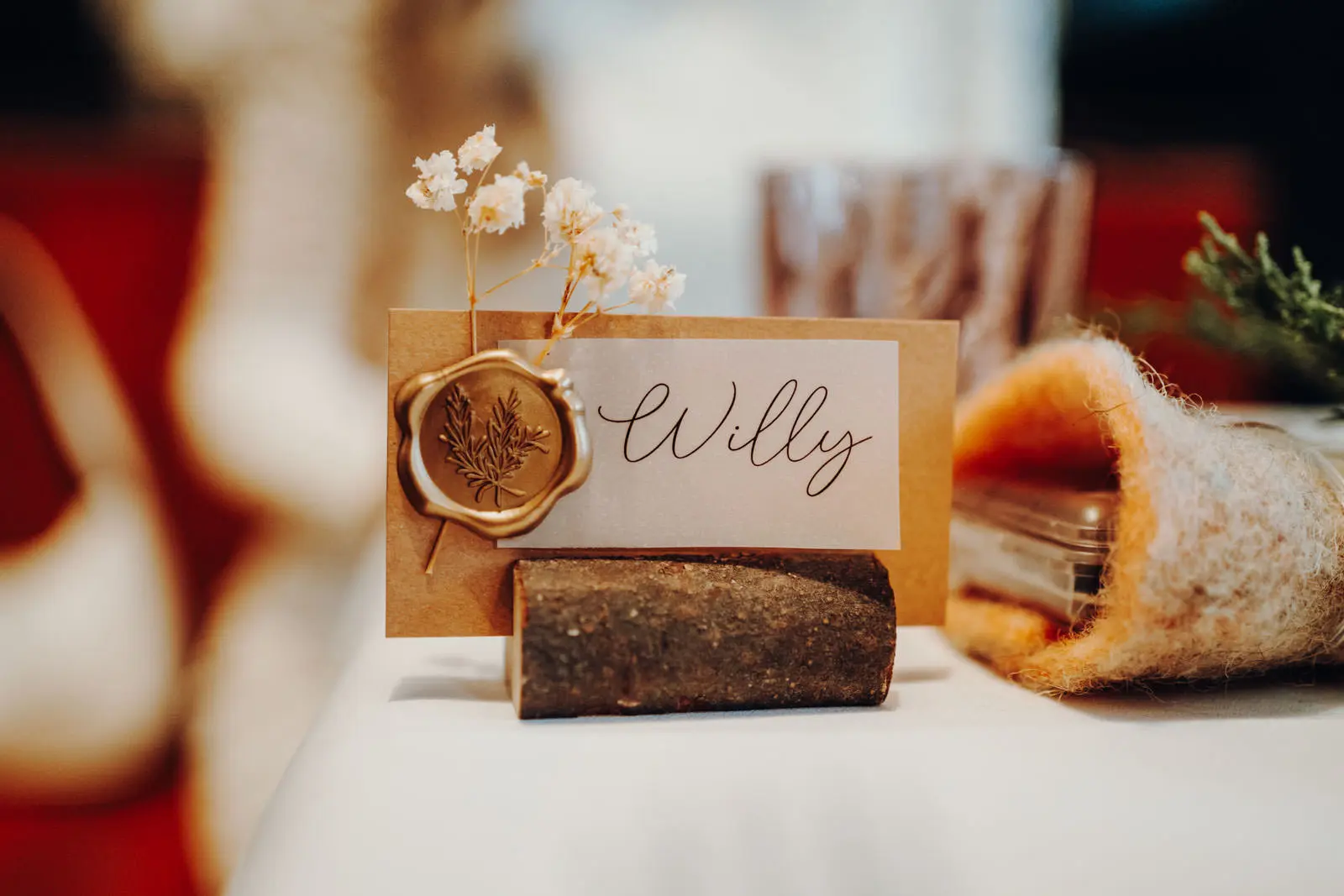 Hochzeitliche Tischkarte mit Wachssiegel und Schleierkraut Personalisierte Tischkarte auf einem Holzstueck, verziert mit einem goldenen Wachssiegel und kleinen Schleierkrautblueten.