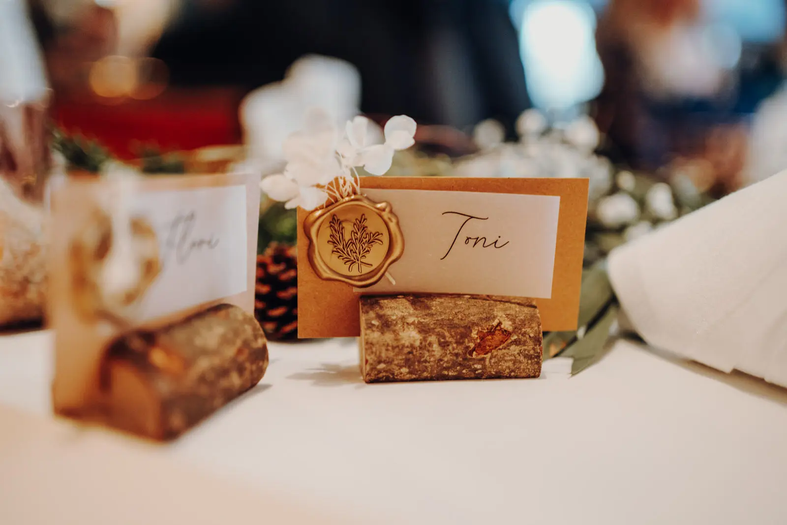 Tischkarten mit Wachssiegel und Beerendeko Nahaufnahme einer Hochzeitstischkarte auf einem Holzstueck, mit goldenem Wachssiegel und winterlicher Beerenzweig-Deko auf dem Tisch.