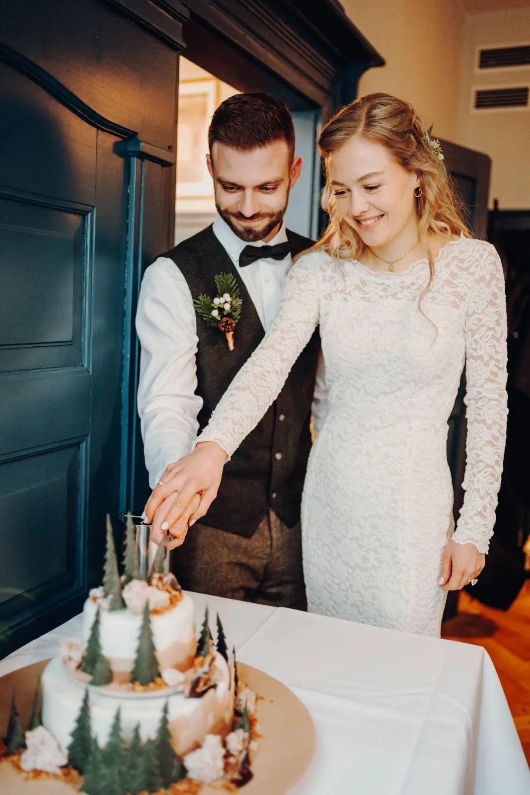 Brautpaar schneidet Hochzeitstorte mit Tannenbaum-Deko an Braut und Braeutigam schneiden gemeinsam die Hochzeitstorte mit winterlicher Tannenbaum-Deko an und laecheln dabei.
